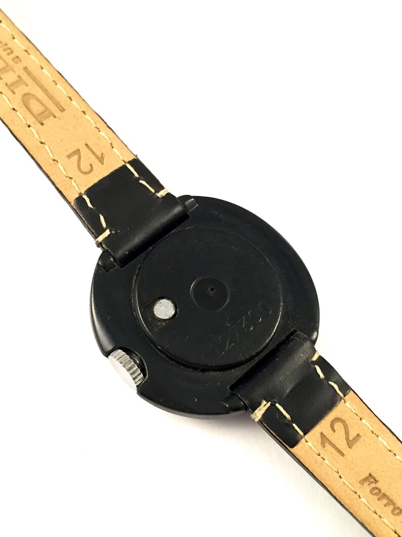 May include: Black round wristwatch with a black leather band. The watch face is covered with a clear plastic cover. The back of the watch has the text "21753" printed on it.
