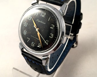 Può includere: Un orologio da polso con quadrante nero, cassa argentata e cinturino in pelle nera con cuciture blu. Il quadrante dell'orologio ha numeri bianchi e una lancetta dei secondi dorata. Il quadrante dell'orologio ha anche il testo "Кировские" stampato su di esso.