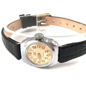 May include: A silver wristwatch with a black leather band. The watch face is cream colored with black Arabic numerals and a gold-colored second hand.