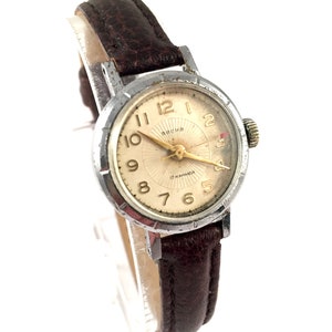 May include: A silver wristwatch with a brown leather strap. The watch face is white with black Roman numeral hour markers and black minute markers. The watch has a gold second hand and the text "ARCHA 17 JEWELS" is printed on the face.