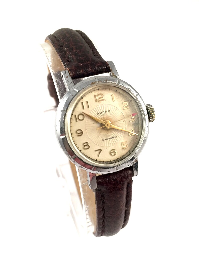 May include: A silver wristwatch with a brown leather strap. The watch face is white with black Roman numeral hour markers and black minute markers. The watch has a gold second hand and the text "ARCHA 17 JEWELS" is printed on the face.