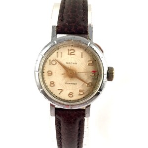 May include: A silver wristwatch with a brown leather strap. The watch face is white with black numbers and a gold-coloured second hand. The watch face also has the text "Весна" and "17 камней".