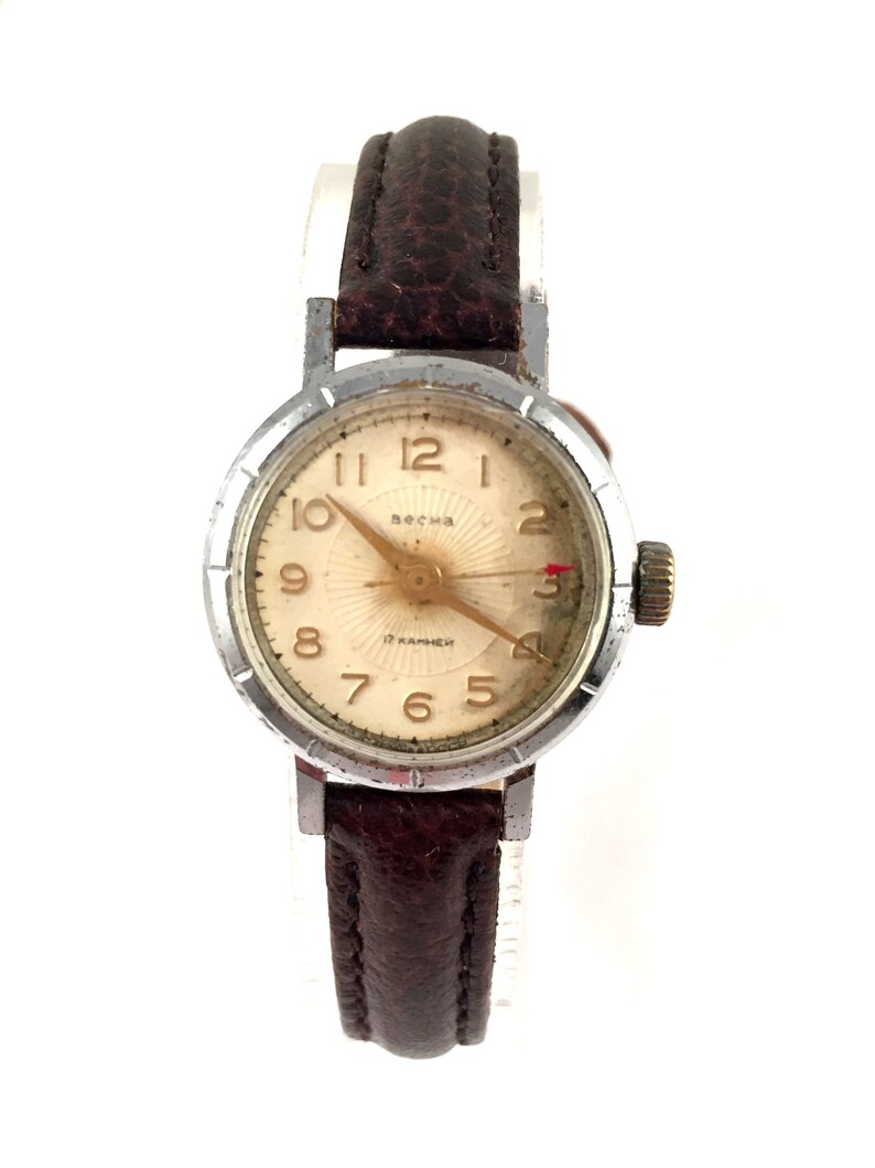 May include: A silver wristwatch with a brown leather strap. The watch face is white with black numbers and a gold-coloured second hand. The watch face also has the text "Весна" and "17 камней".