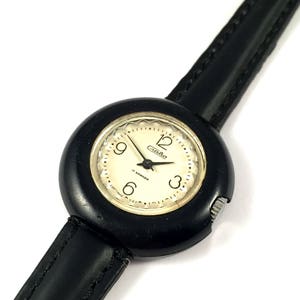 May include: A black wristwatch with a round, black face and a black leather band. The watch face has white numbers and hands, and the word "Rada" is printed in gold letters.
