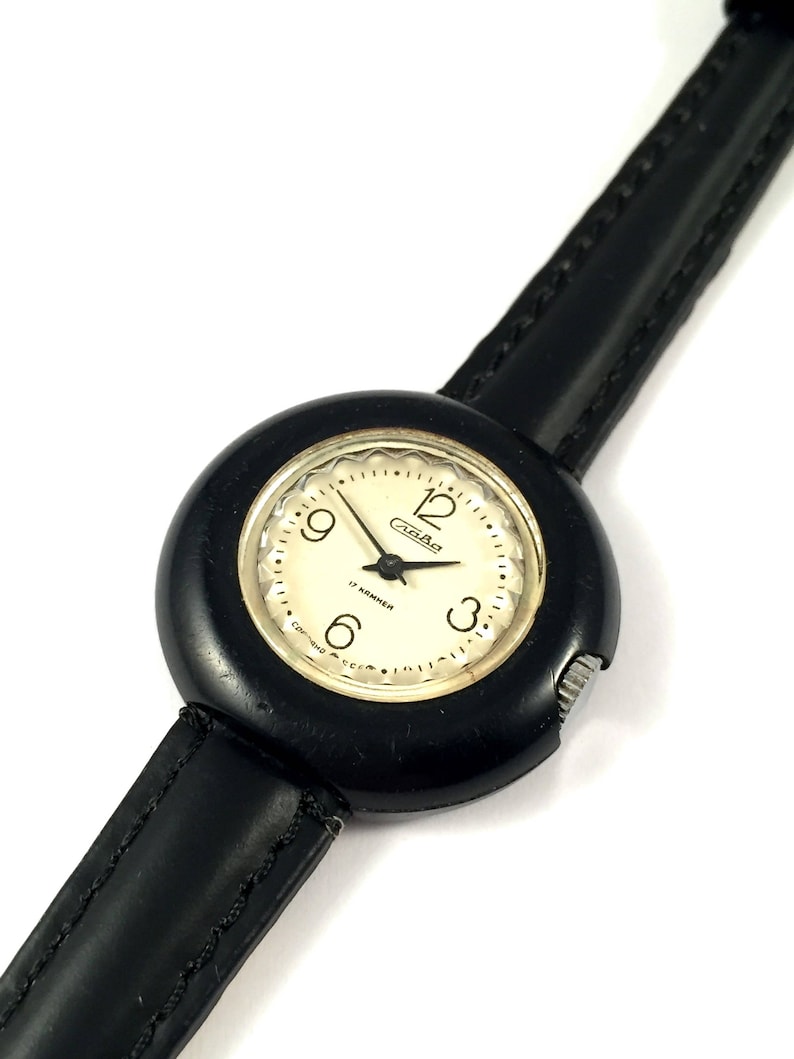 May include: A black wristwatch with a round, black face and a black leather band. The watch face has white numbers and hands, and the word "Rada" is printed in gold letters.