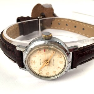 May include: A silver wristwatch with a white face and black leather strap. The watch face has black Roman numeral hour markers and a gold second hand. The watch face also has the word "СССР" printed on it.