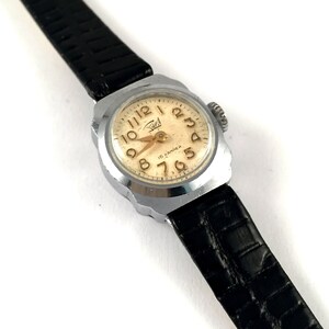 May include: A silver wristwatch with a black leather band. The watch face is white with black Roman numeral hour markers and black hands. The watch face also has the text "16 KAMHEH" in gold lettering.