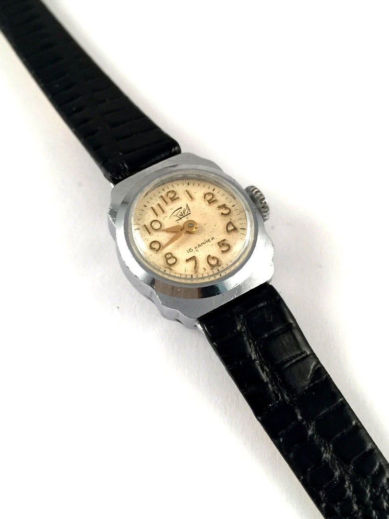 May include: A silver wristwatch with a black leather band. The watch face is white with black Roman numeral hour markers and black hands. The watch face also has the text "16 KAMHEH" in gold lettering.