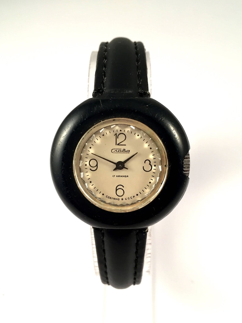 May include: A black wristwatch with a round, black face and a black leather band. The watch face has white numbers and hands, and the text "Слава 17 камней Сделано в СССР" is printed on the face.