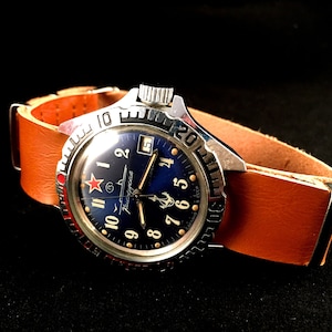 May include: A silver wristwatch with a blue face and red accents. The watch has a brown leather strap and a date window at the 3 o'clock position. The watch face features the text "Poljot" and an anchor symbol.