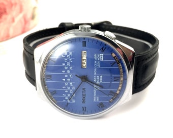 Puede incluir: Un reloj de pulsera plateado con una esfera azul y una correa de cuero negro. La esfera del reloj presenta un calendario con los días de la semana y los meses del año en escritura cirílica. La esfera del reloj también incluye el texto "PAKETA" y "CPA 5".