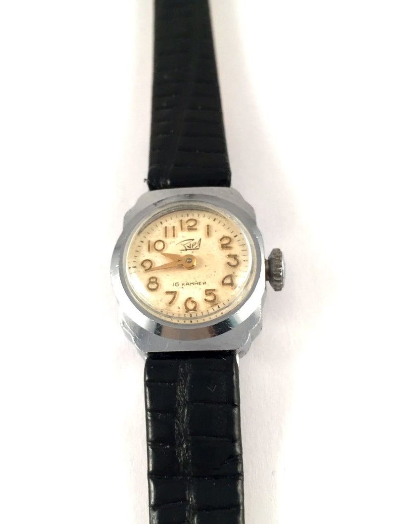 May include: A silver wristwatch with a white face and black leather band. The watch face has black Roman numeral hour markers and gold Arabic numeral minute markers. The watch face also has the text "16 камней" in gold lettering.