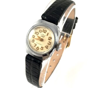 May include: A silver wristwatch with a black leather band. The watch face is white with black Roman numeral hour markers and black Arabic numeral minute markers. The watch has a gold-colored second hand and the text "15 KAMEX" is printed on the watch face.