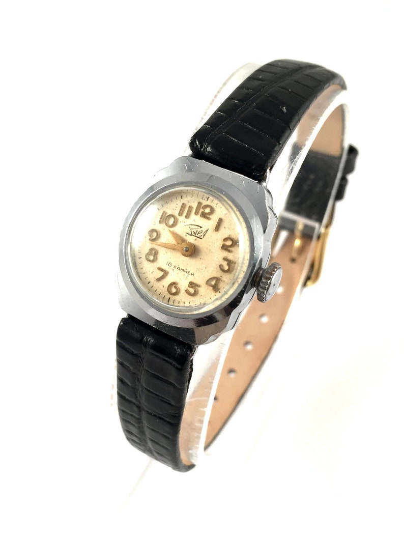 May include: A silver wristwatch with a black leather band. The watch face is white with black Roman numeral hour markers and black Arabic numeral minute markers. The watch has a gold-colored second hand and the text "15 KAMEX" is printed on the watch face.
