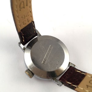 May include: A silver wristwatch with a brown leather strap. The watch face is covered with a clear glass. The back of the watch has the text "Shockproof" engraved on it.