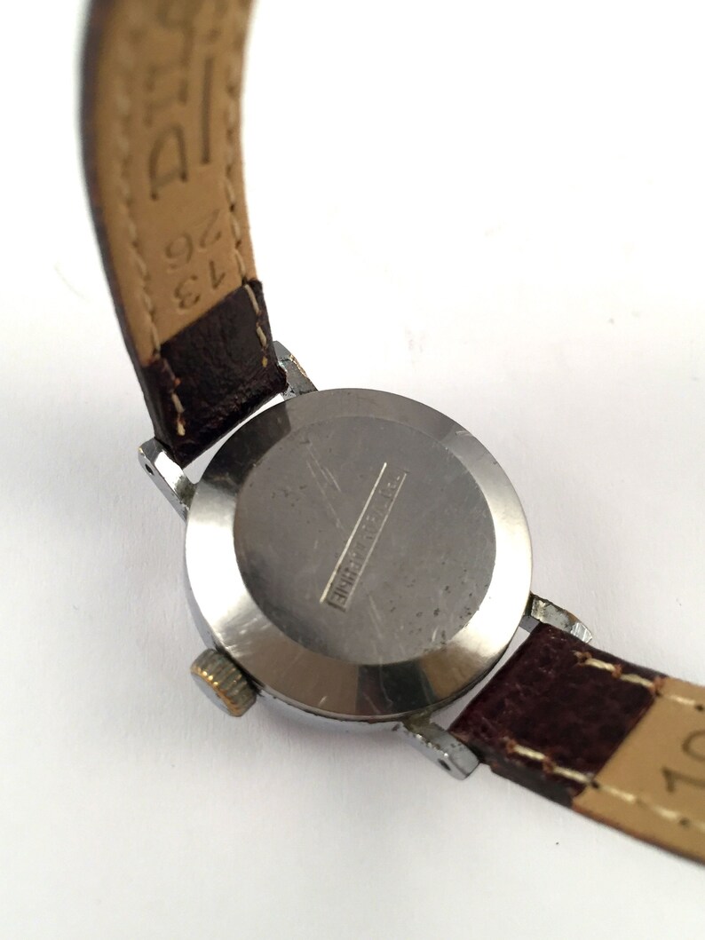 May include: A silver wristwatch with a brown leather strap. The watch face is covered with a clear glass. The back of the watch has the text "Shockproof" engraved on it.