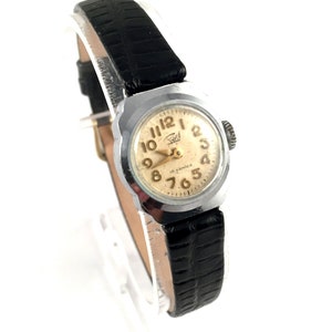 May include: A silver-toned wristwatch with a black leather band. The watch face is white with black Arabic numerals and a gold-colored second hand. The watch has a square case and a small crown on the right side.