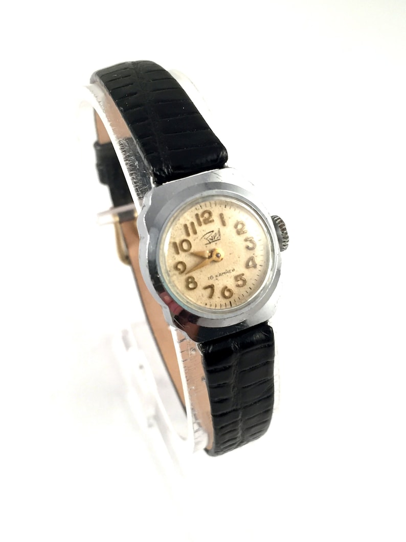 May include: A silver-toned wristwatch with a black leather band. The watch face is white with black Arabic numerals and a gold-colored second hand. The watch has a square case and a small crown on the right side.