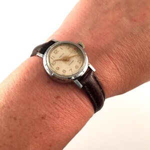 May include: A silver wristwatch with a brown leather strap. The watch face is white with black numbers and a red second hand. The watch has the word "Breitling" printed on the face.