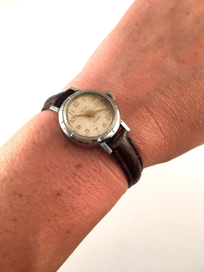 May include: A silver wristwatch with a brown leather strap. The watch face is white with black numbers and a red second hand. The watch has the word "Breitling" printed on the face.