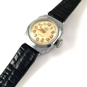 May include: A silver wristwatch with a black leather band. The watch face is white with black Roman numeral hour markers and gold Arabic numeral minute markers. The watch face also has the text "15 камней" in Cyrillic script.