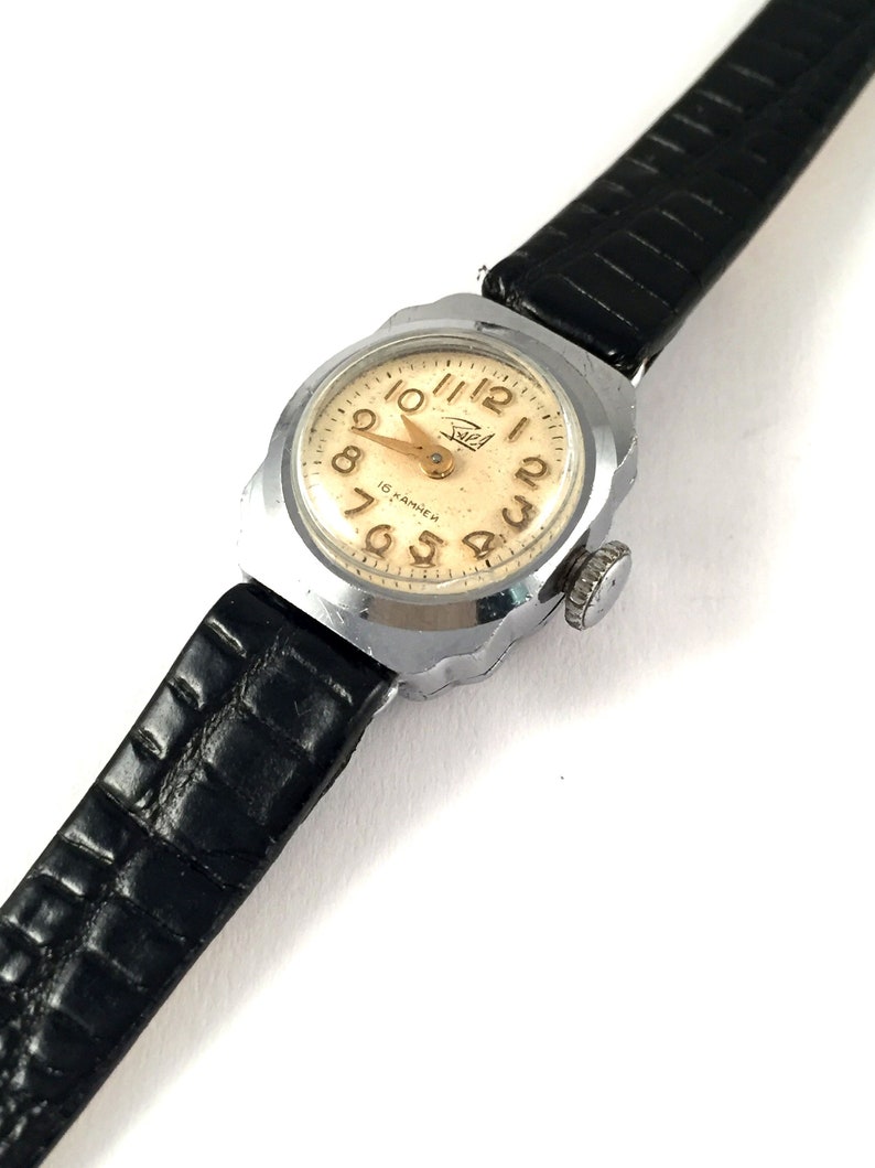 May include: A silver wristwatch with a black leather band. The watch face is white with black Roman numeral hour markers and gold Arabic numeral minute markers. The watch face also has the text "15 камней" in Cyrillic script.