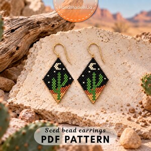 May include: A pair of diamond-shaped seed bead earrings featuring a desert night scene with a green cactus, a white crescent moon, and small white stars against a black background, finished with gold-toned metal ear wires for pierced ears.