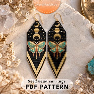 May include: A pair of long, diamond-shaped seed bead earrings featuring a black background with a gold moon phase design at the top and a teal and gold moth graphic in the center. The bottom edges are finished with gold and black fringe.