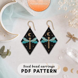 May include: Diamond-shaped seed bead earrings featuring a black background with a central dragonfly motif in turquoise and gold. The design includes a small gold crescent moon above the dragonfly. The earrings hang from gold-toned metal hooks.