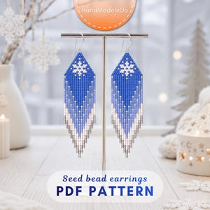May include: A pair of long, diamond-shaped seed bead earrings featuring a blue and white gradient pattern with a white snowflake design at the top. The earrings hang from silver-toned hooks and display a fringe-like bottom edge.