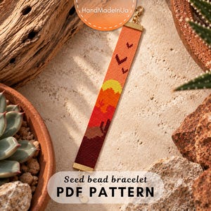May include: A rectangular seed bead bracelet featuring a desert landscape pattern with a yellow sun, orange sky, and dark red ground. The design includes small bird silhouettes and cactus shapes. It has gold-toned metal end caps and a lobster clasp.