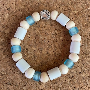 May include: A beaded bracelet featuring a paw print charm. The bracelet is made with light blue, white, and wooden beads. The paw print charm is silver-colored. The bracelet is displayed on a brown, textured surface.