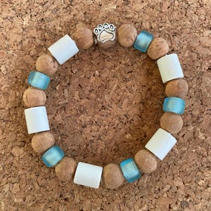 Op de afbeelding: Een armband met een bedel in de vorm van een pootafdruk. De armband bevat lichtbruine, ronde kralen, witte rechthoekige kralen en doorschijnende blauwe vierkante kralen. De bedel is zilverkleurig met een pootafdruk.