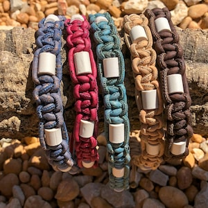 May include: Five different colored braided dog collars with white ceramic beads. The collars are made of cork and are shown on a wooden surface.