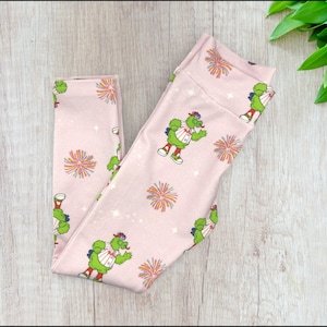 May include: Pink leggings with a green cartoon character wearing a baseball uniform and holding a bat. The leggings have a white background with a repeating pattern of fireworks.