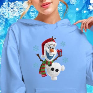 May include: Light blue hoodie featuring a festive Olaf design. The embroidered Olaf wears a Santa hat and a striped scarf, holding a gift. The hoodie has a front pocket and a drawstring hood, perfect for winter wear.