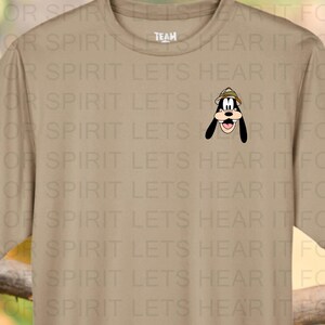 May include: Desert khaki t-shirt featuring a small embroidered image of Goofy wearing a safari hat. The shirt is displayed with a jaguar and costumed Mickey and Minnie Mouse characters. The text "Shown on Desert Khaki" is at the bottom.