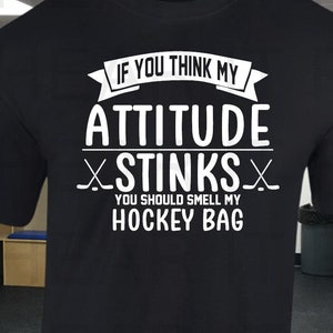 May include: A black t-shirt with white text that reads 'If you think my attitude stinks you should smell my hockey bag.' The t-shirt is displayed in a hockey locker room with hockey equipment on the floor.