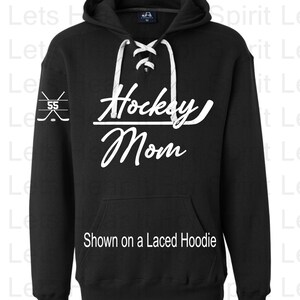K&ouml;nnte beinhalten: Schwarzer Hoodie mit einem wei&szlig;en Schn&uuml;rdetail am Ausschnitt. Der Hoodie hat eine wei&szlig;e Grafik mit der Aufschrift "Hockey Mom" und einem Hockeystock- und Puck-Design am linken &Auml;rmel.