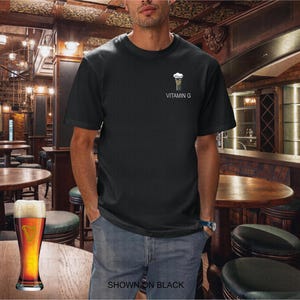 K&ouml;nnte beinhalten: Schwarzes T-Shirt mit einer kleinen Grafik eines Bierglases und dem Text "VITAMIN G". Das Bild zeigt einen Mann in einer Bar, mit einem Bier auf dem Tisch. Der Text "SHOWN ON BLACK" steht unten.