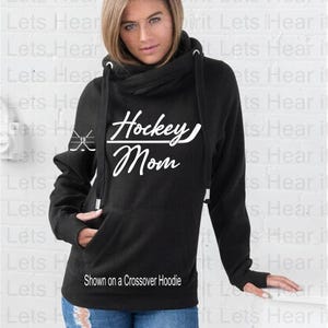 K&ouml;nnte beinhalten: Schwarzer Crossover-Hoodie mit einem wei&szlig;en Aufdruck, der "Hockey Mom" mit einem Hockey-Schl&auml;ger- und Puck-Design zeigt. Der Hoodie wird an einem Model gezeigt.