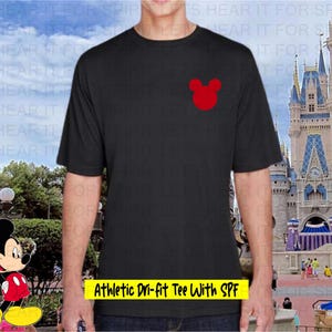 May include: Black athletic t-shirt with a red Mickey Mouse head logo on the chest. The shirt has short sleeves and the text "Athletic Dri-fit Tee With SPF" in a yellow box. The background features a castle and cartoon character.