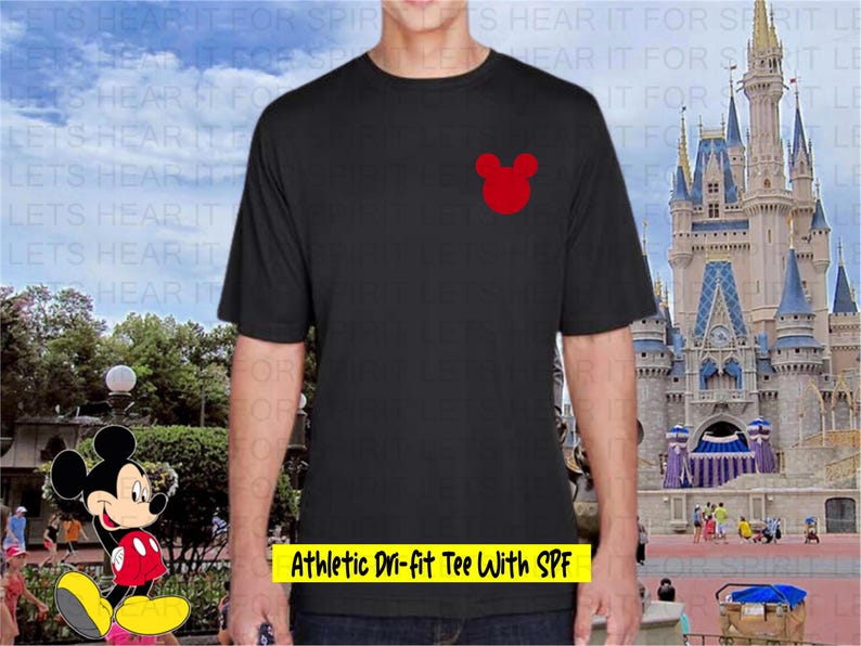 May include: Black athletic t-shirt with a red Mickey Mouse head logo on the chest. The shirt has short sleeves and the text "Athletic Dri-fit Tee With SPF" in a yellow box. The background features a castle and cartoon character.