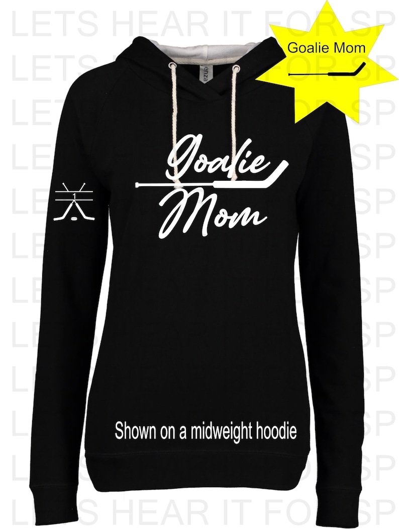 Puede incluir: Sudadera con capucha negra con un gr&aacute;fico blanco que dice "Goalie Mom" y un palo de hockey. La sudadera se muestra en una modelo.