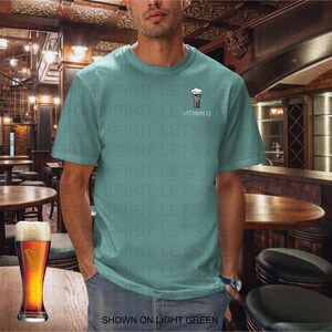 K&ouml;nnte beinhalten: Hellgr&uuml;nes T-Shirt mit einer kleinen Bierglas-Grafik und dem Text "Vitamin G". Ein Bierkrug steht auf einem Holztisch in einem Pub. Der Text "SHOWN ON LIGHT GREEN" steht unten.