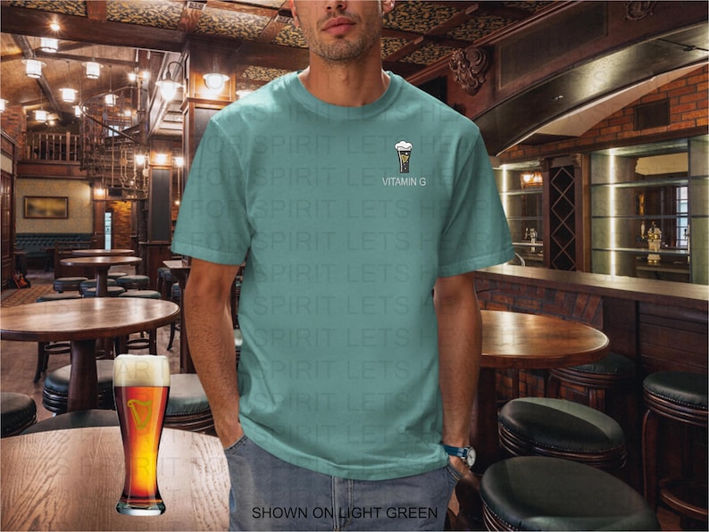 K&ouml;nnte beinhalten: Hellgr&uuml;nes T-Shirt mit einer kleinen Bierglas-Grafik und dem Text "Vitamin G". Ein Bierkrug steht auf einem Holztisch in einem Pub. Der Text "SHOWN ON LIGHT GREEN" steht unten.