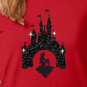 Puede incluir: Camiseta roja con una silueta negra brillante de un castillo y una sirena. El castillo tiene m&uacute;ltiples torres y banderas. La sirena est&aacute; sentada en una roca, con un reflejo en el agua. El dise&ntilde;o est&aacute; sobre un fondo rojo.