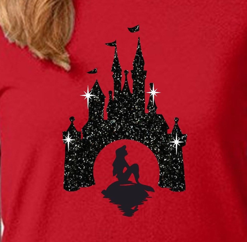 Puede incluir: Camiseta roja con una silueta negra brillante de un castillo y una sirena. El castillo tiene m&uacute;ltiples torres y banderas. La sirena est&aacute; sentada en una roca, con un reflejo en el agua. El dise&ntilde;o est&aacute; sobre un fondo rojo.