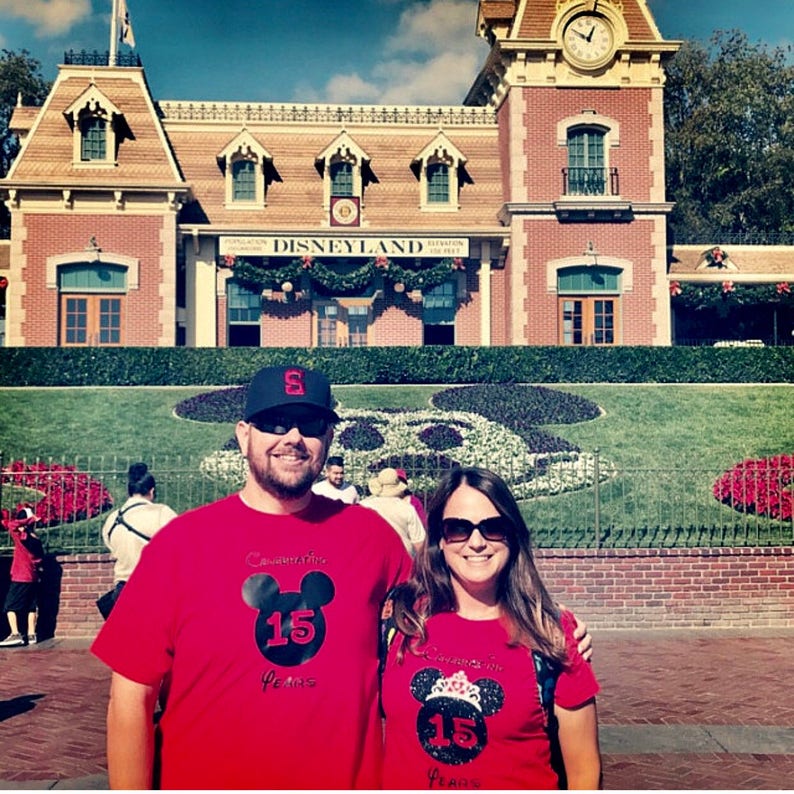 May include: A couple wearing red t-shirts with a black Mickey Mouse head and the text "Celebrating 15 Years" in front of the Disneyland entrance.