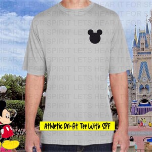 May include: A light gray athletic t-shirt with a black Mickey Mouse head silhouette on the chest. The text "Athletic Dri-fit Tee With SPF" is in a yellow rectangle at the bottom. The background shows a castle and a cartoon character.